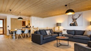 Ein modernes Wohnzimmer mit Holzboden, einer großen Sofagarnitur, einem Essbereich mit Holztisch und Stühlen sowie einer Küche im Hintergrund.