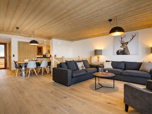 A modern living room with a wooden ceiling, a large sofa set, a dining area with a wooden table and chairs, and a kitchen in the background.