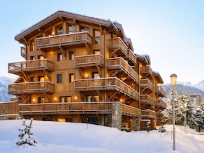 A multi-story wooden lodge with balconies and a stone foundation, surrounded by snow and mountains.