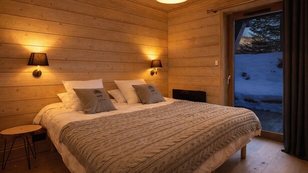 Ein gemütliches Schlafzimmer in einer Holzhütte mit einem großen Bett, zwei Wandleuchten und Blick auf eine verschneite Landschaft durch eine Glastür.