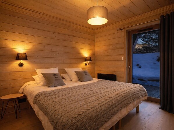 Un dormitorio acogedor con cabaña de madera, una cama grande, dos lámparas fijadas a la pared y vistas a un paisaje nevado a través de un ventanal.