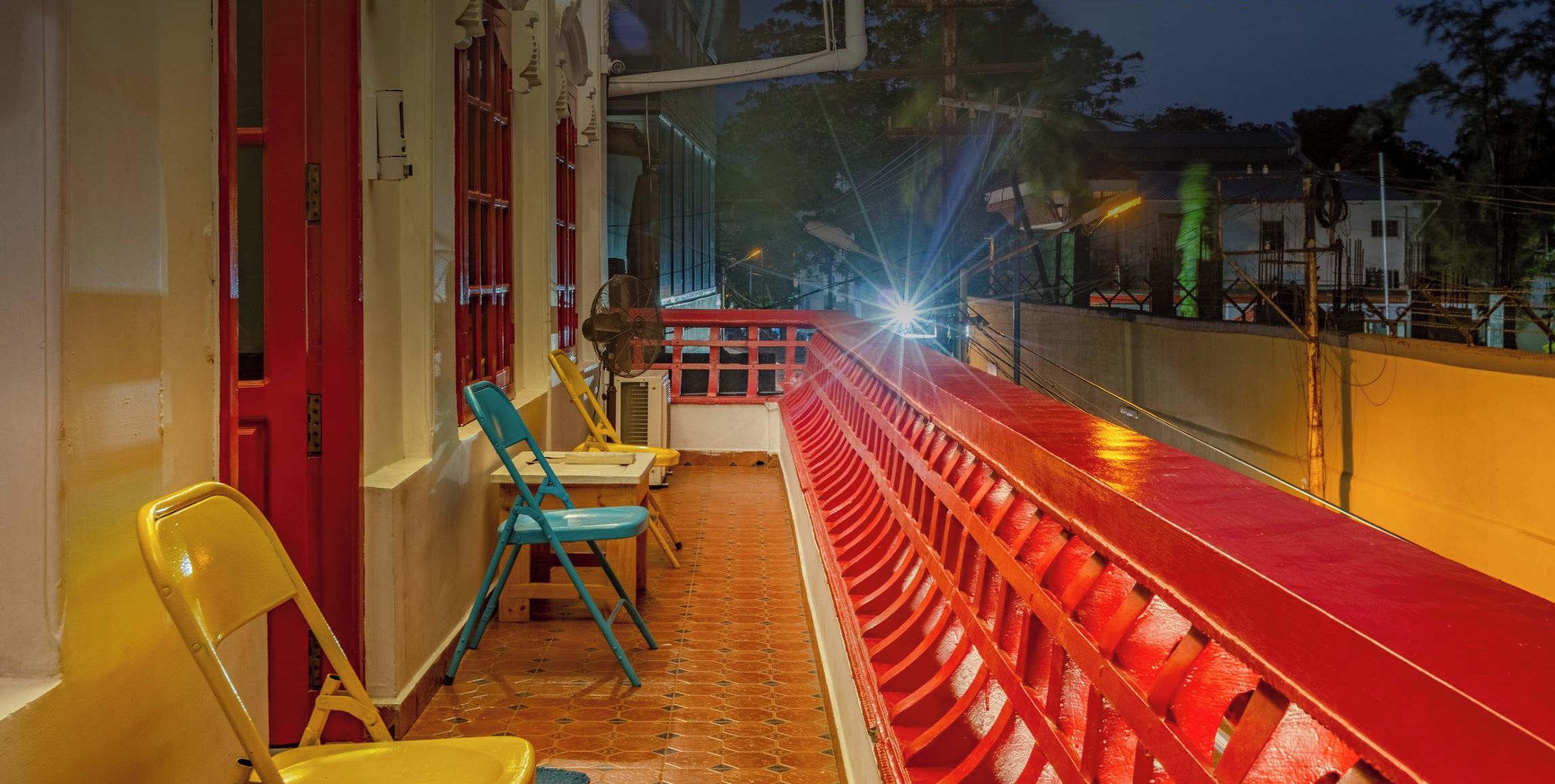 A balcony with a red railing, chairs, and a view of the street at night.
