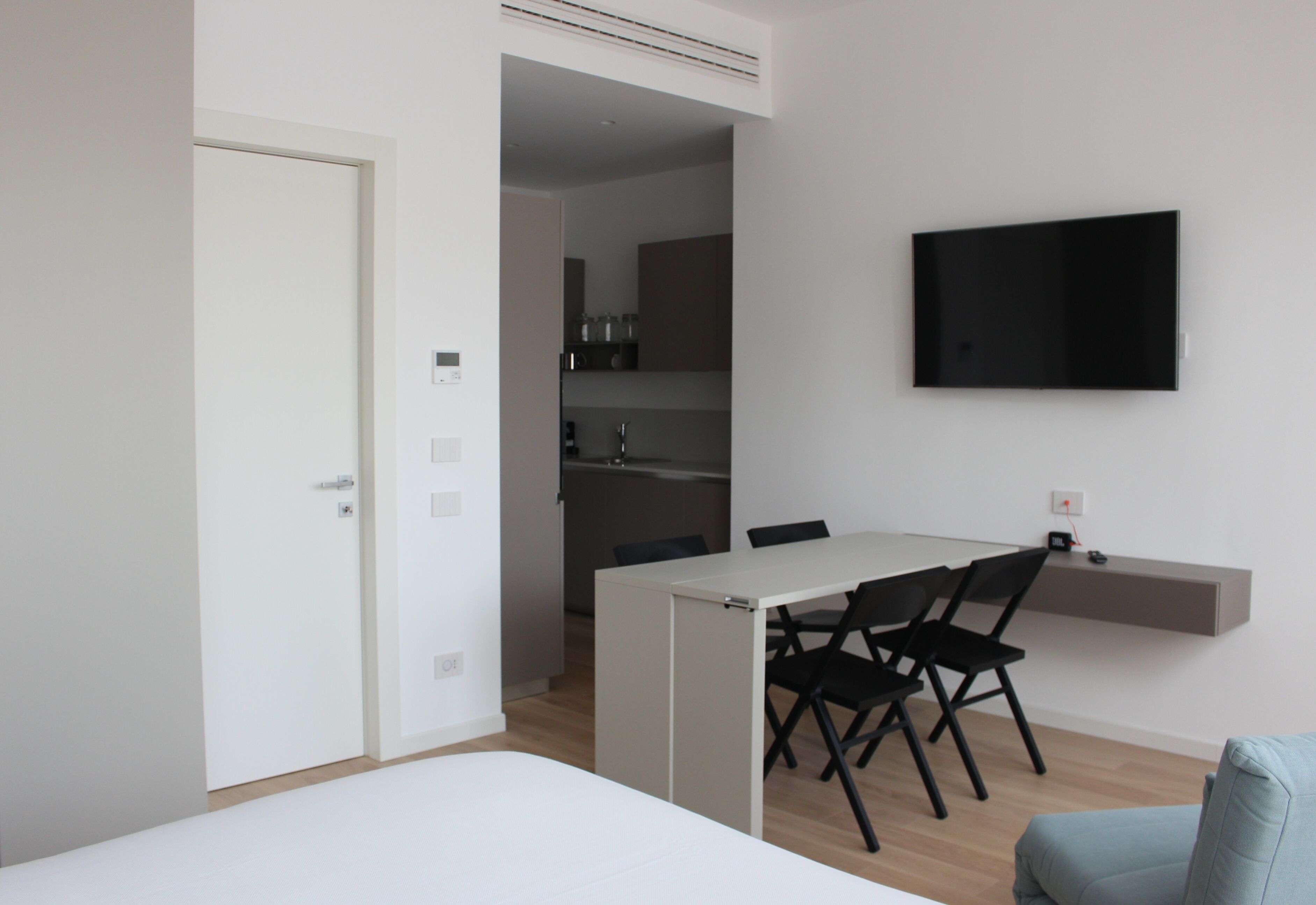 Een moderne, minimalistische kamer met een bed, een eettafel met stoelen, een aan de muur bevestigde televisie en een zichtbare keuken door een open deur.