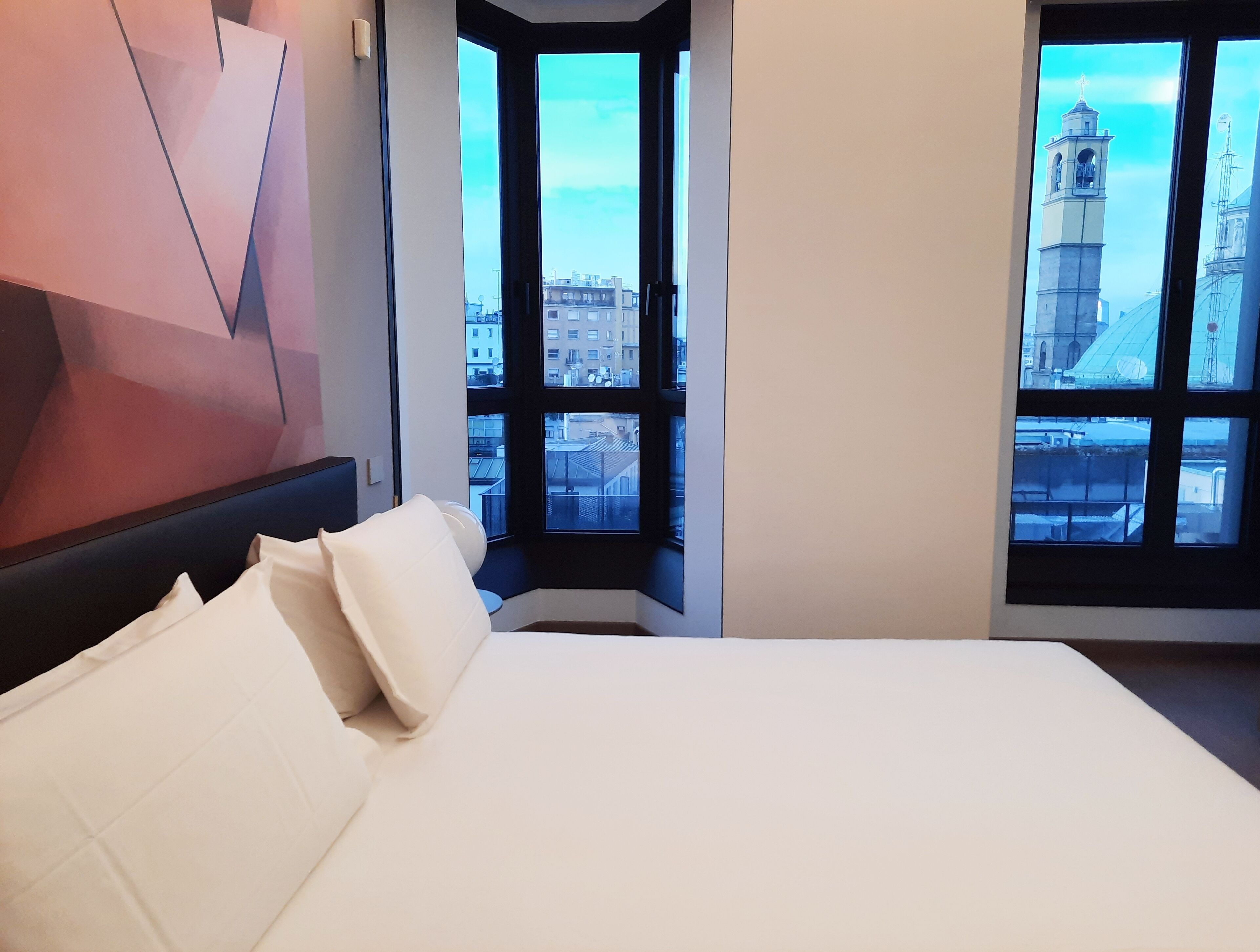 A modern hotel room with a large bed, a geometric wall art piece, and a view of a cityscape through large windows.