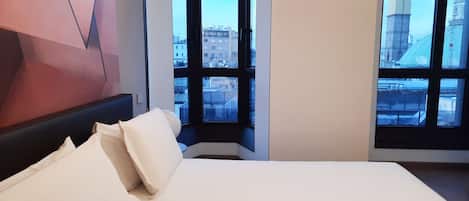 Habitación de hotel moderna con una cama grande, una obra de arte geométrica en la pared y vistas a un paisaje urbano a través de amplios ventanales.