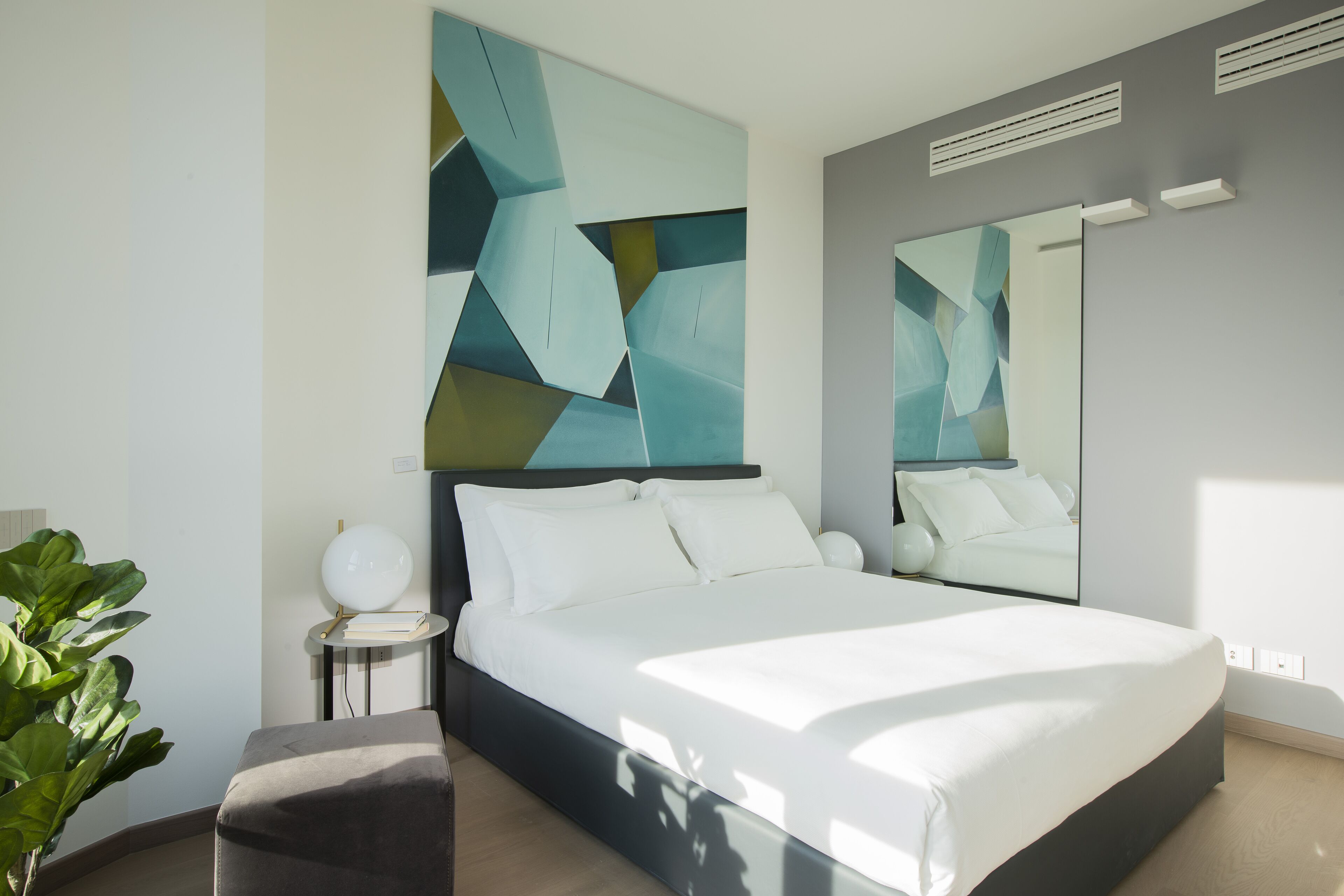 A modern bedroom with a large bed, a bedside table, a mirror, and abstract wall art.