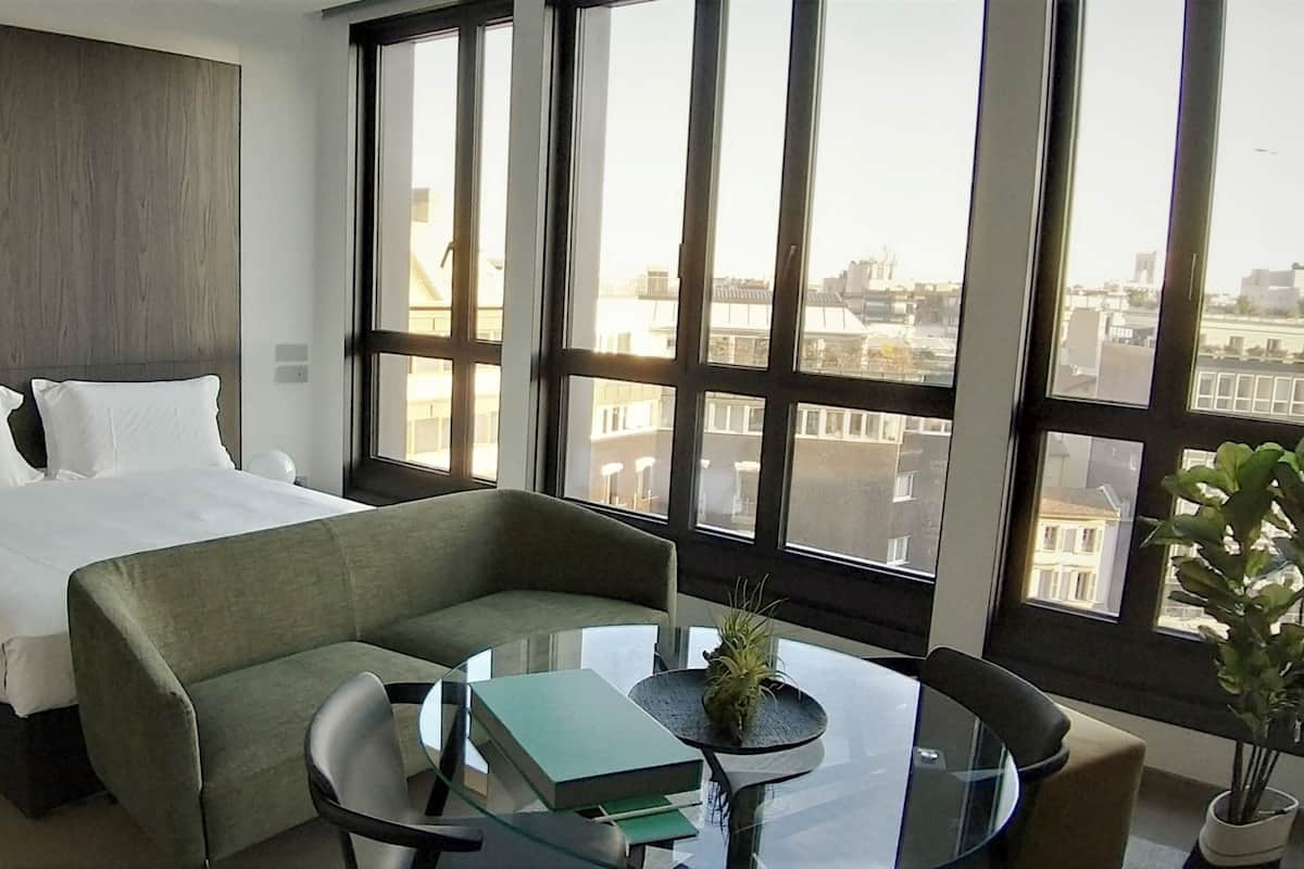 Une chambre d’hôtel avec un lit, un canapé, une table en verre ornée d’une plante, et une vue sur la ville grâce à de grandes fenêtres.