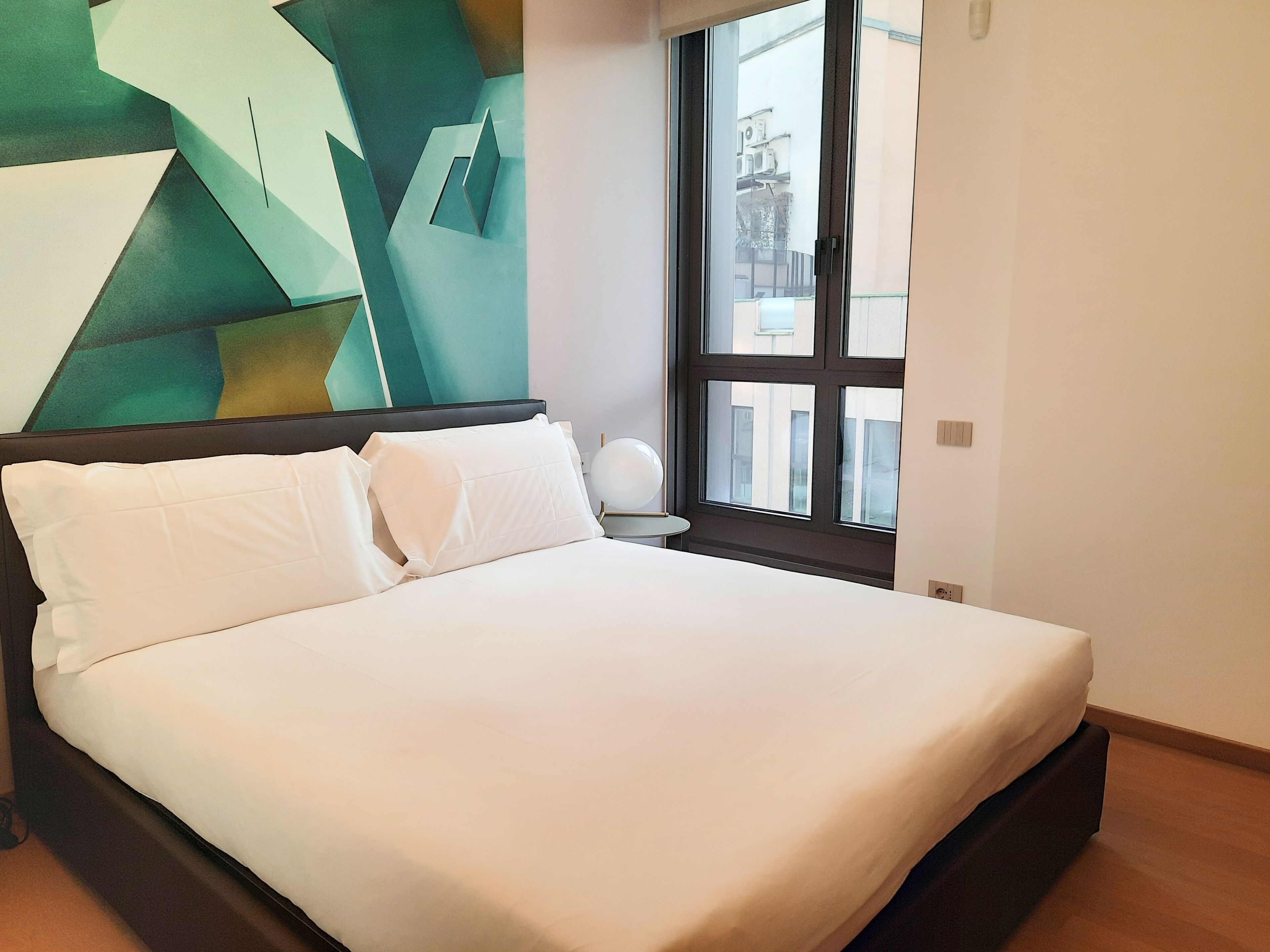 Un lit bien fait, avec du linge de lit blanc, dans une chambre moderne agrémentée d’une œuvre d’art géométrique au mur et d’une grande fenêtre.
