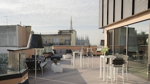 Una terraza en la azotea con asientos al aire libre, vista al horizonte de la ciudad y fachada de un edificio moderno.