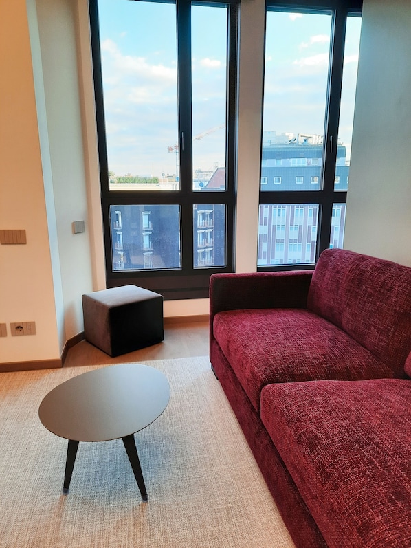 Una sala de estar moderna con un sofá rojo, una pequeña mesa redonda y vistas a edificios a través de amplios ventanales.