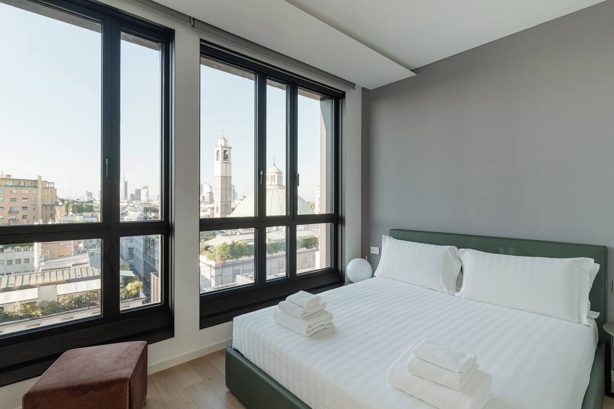 Une chambre avec une grande fenêtre, un lit recouvert de draps blancs, une tête de lit verte et un petit pouf marron.
