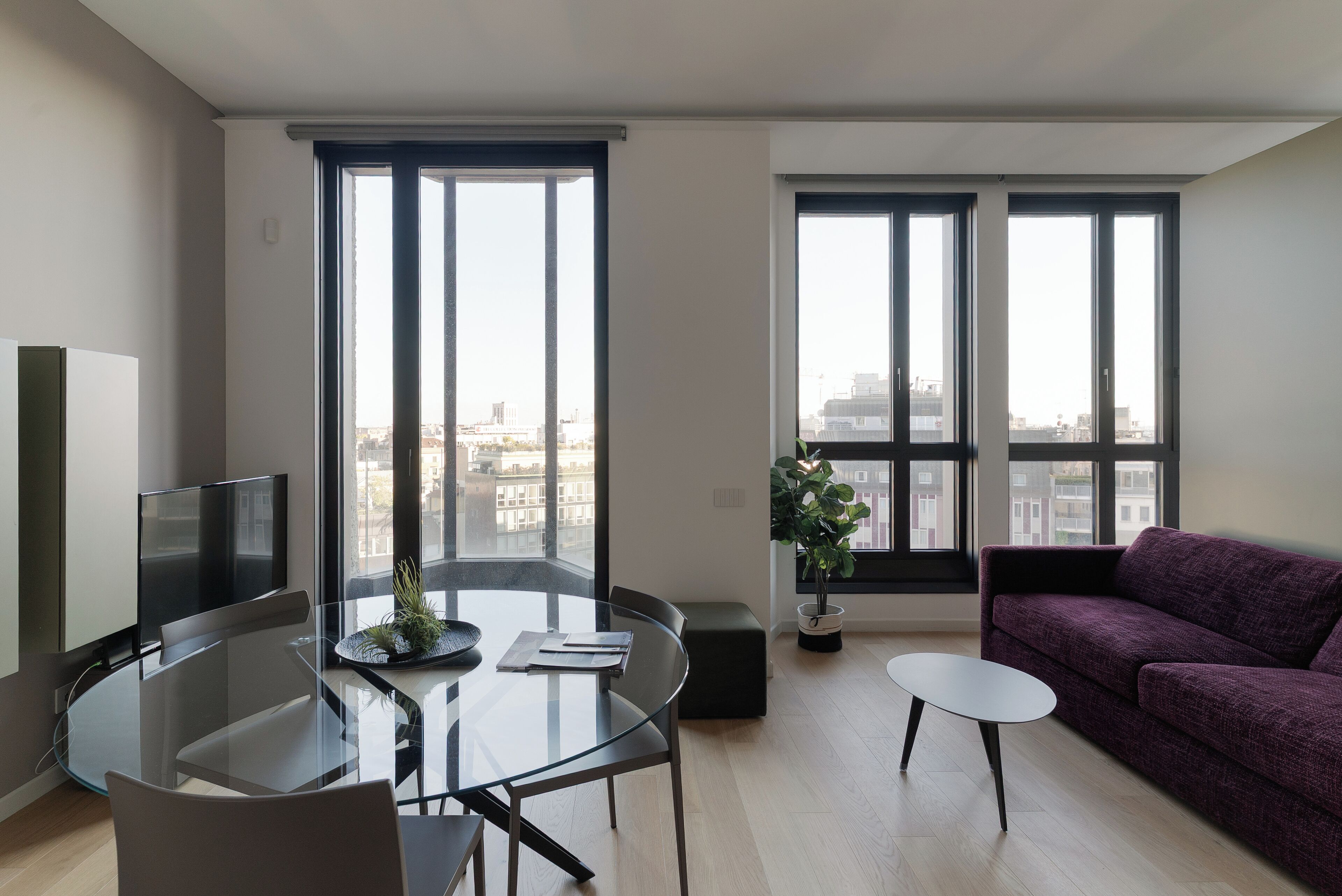 Un salon moderne avec une table à manger en verre, un canapé et de grandes fenêtres donnant sur la ville.