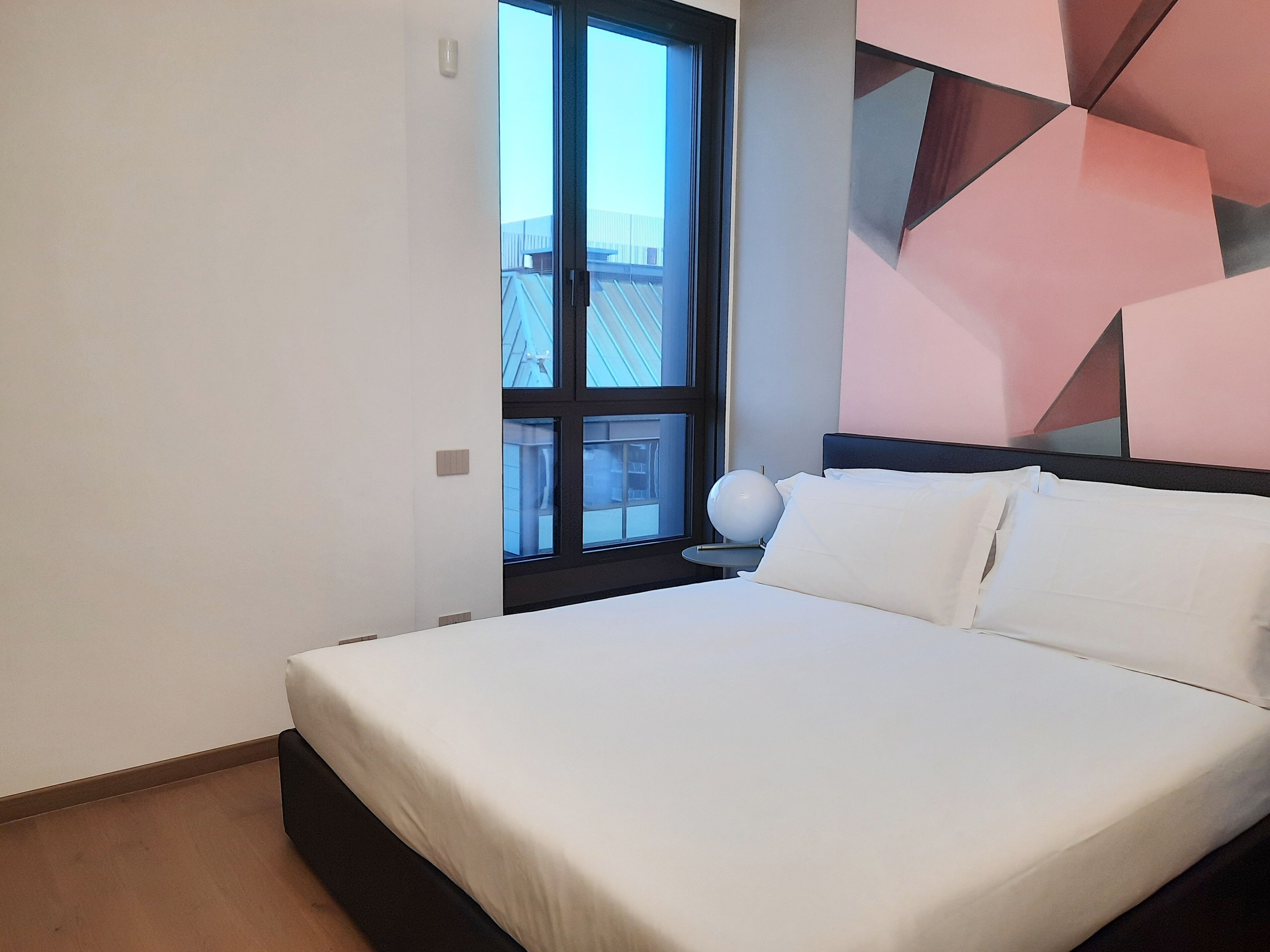 Ein modernes Hotelzimmer mit einem großen Bett, einem Nachttisch mit Lampe und Blick auf Gebäude durch ein Fenster.