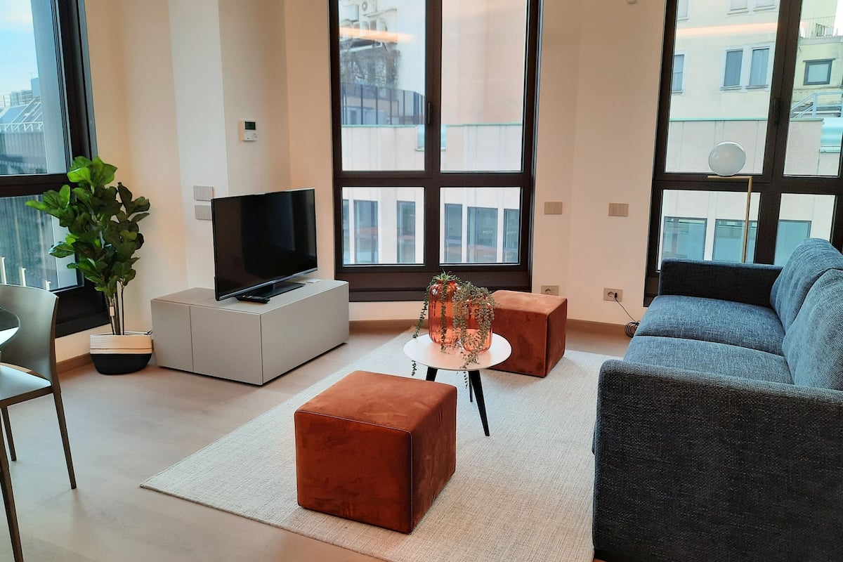 Un salon moderne avec un téléviseur à écran plat, une plante en pot, une table basse, un canapé et un petit pouf.