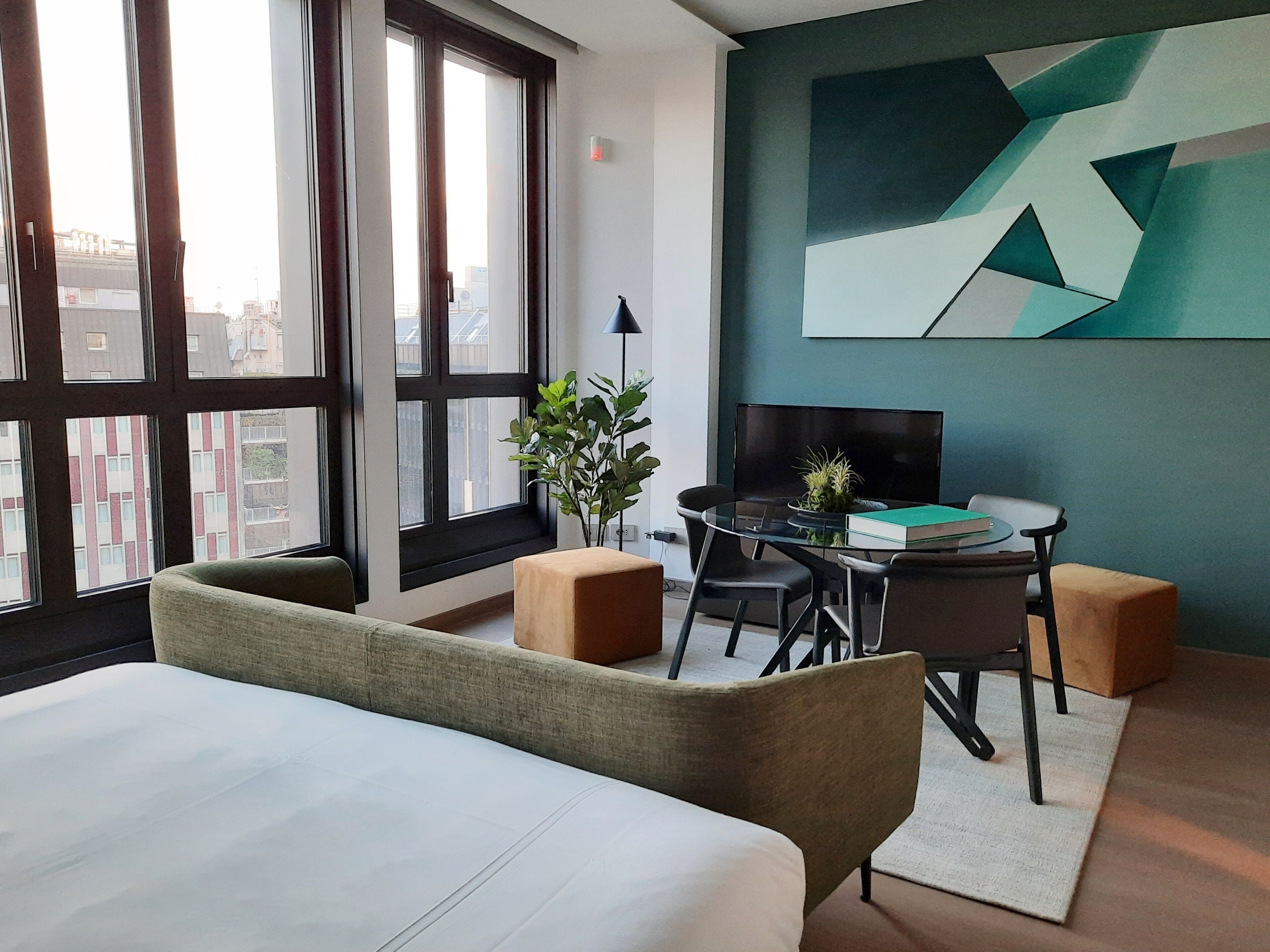 A modern hotel room with a large bed, a desk with a chair, a small table with a plant, and a large window offering a city view.