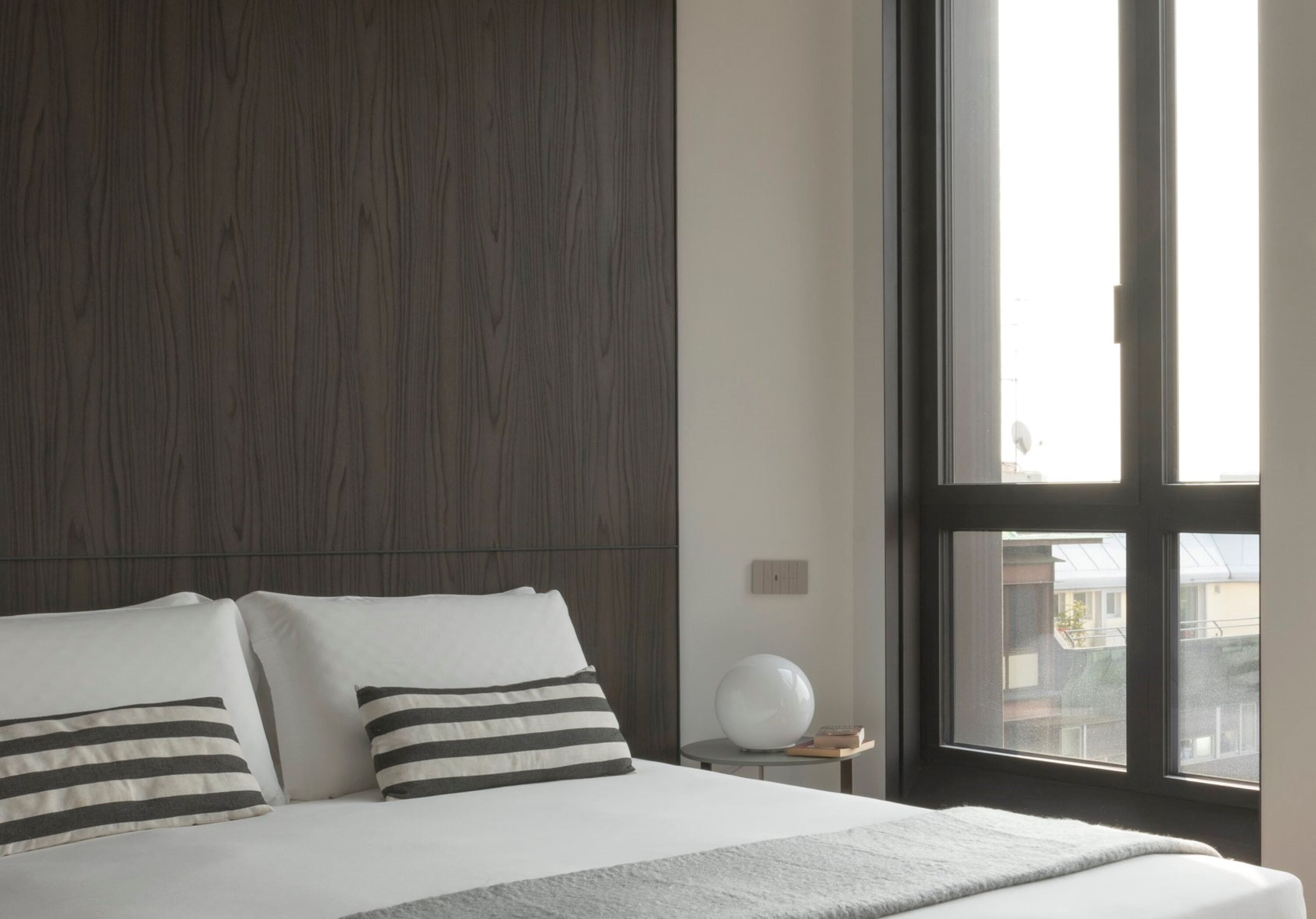 A modern bedroom with a large window, a bed with striped pillows, and a round white table.