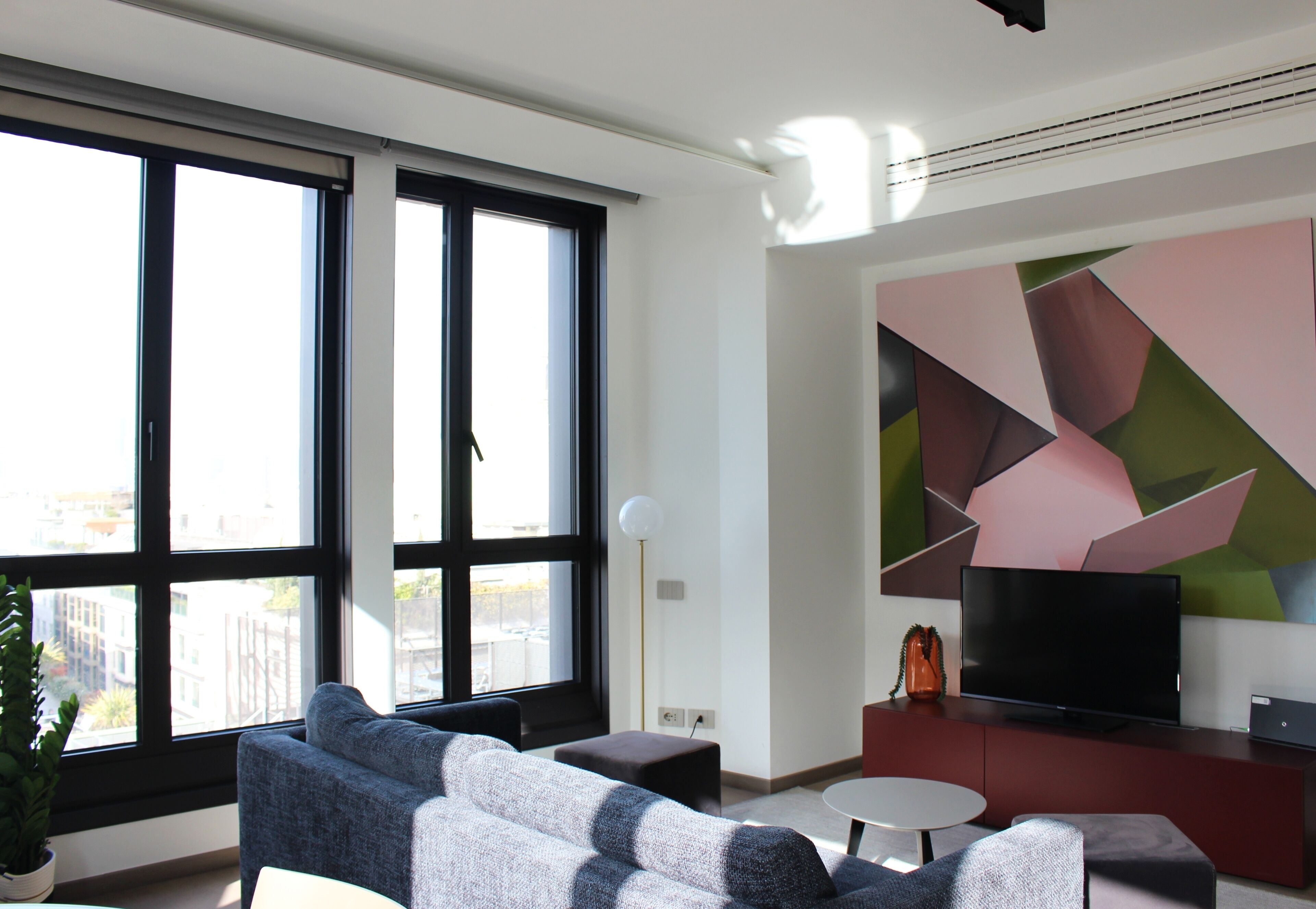 A modern living room with large windows, a flat-screen TV, a red entertainment unit, and a geometric wall art piece.
