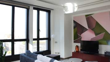 Ein modernes Wohnzimmer mit großen Fenstern, einem Flachbildfernseher, einer roten Unterhaltungsecke und einem Wandbild mit geometrischen Formen.