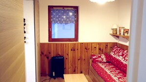 Ett litet rum med trägolv, en säng med rött sänglinne, en träbänk, ett litet bord, ett fönster med gardin och en hängande lampa.