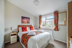 A bedroom with a bed, a nightstand, a lamp, a window with curtains, a painting on the wall, and a radiator. - 2 Bed Cabin - Sleeps 4 - Pets - 5 min to Beach (Bridlington)
