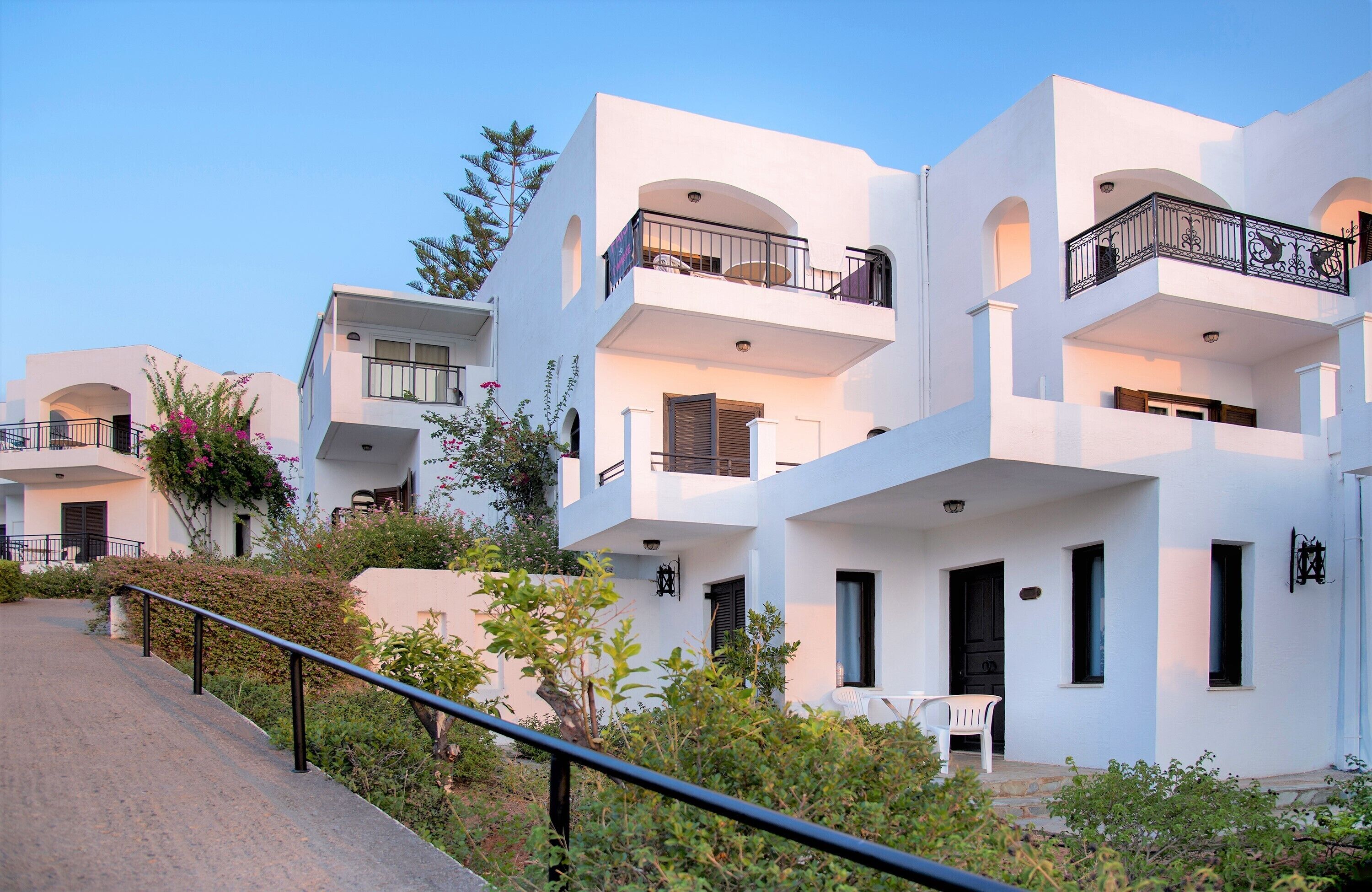 A modern white building with multiple balconies, a paved pathway, and greenery.