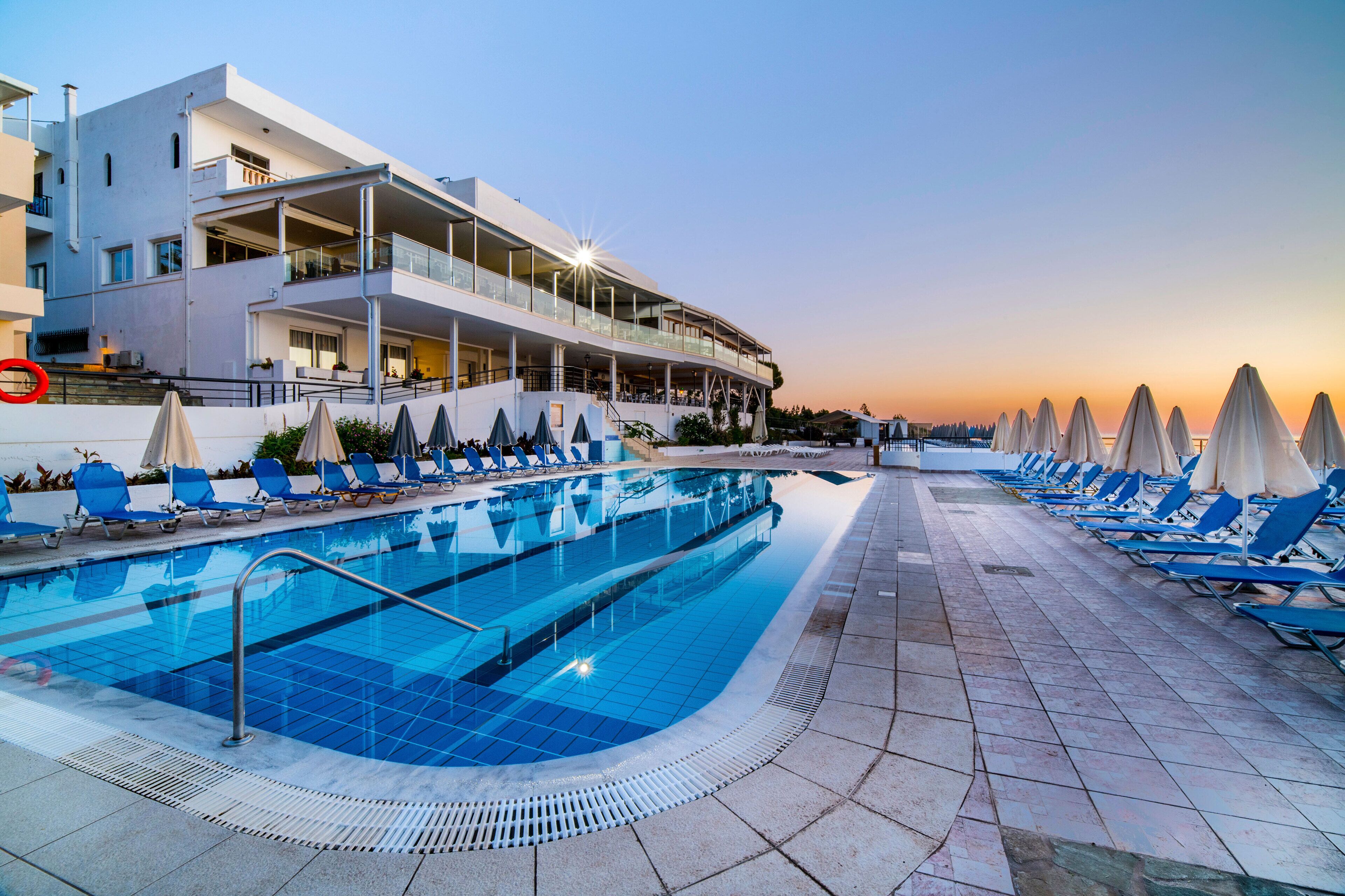 Una piscina con sillas de descanso y sombrillas, un edificio moderno con balcones, y una puesta de sol al fondo.