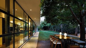 Un espace de restauration extérieur moderne, avec une table et des chaises en bois, un mur de verre d’un côté et une pelouse bien entretenue.