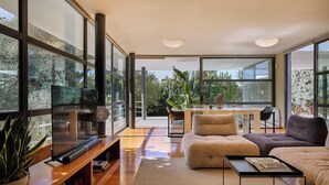 Un salon moderne avec un canapé, une table basse et un téléviseur. Il y a également un coin repas avec une table et des chaises. De grandes fenêtres offrent une vue sur l’extérieur.