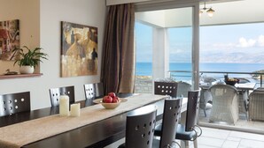 Een eethoek met uitzicht op zee, een groot schilderij aan de muur en een schaal fruit op tafel.