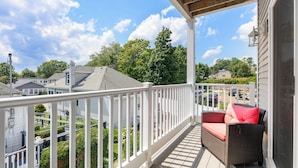 A covered porch with a white railing, a wicker sofa with red cushions, and a view of neighboring houses and trees.