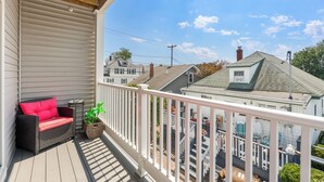A covered porch with a wicker sofa and a potted plant, overlooking a residential area with houses and a clear sky.