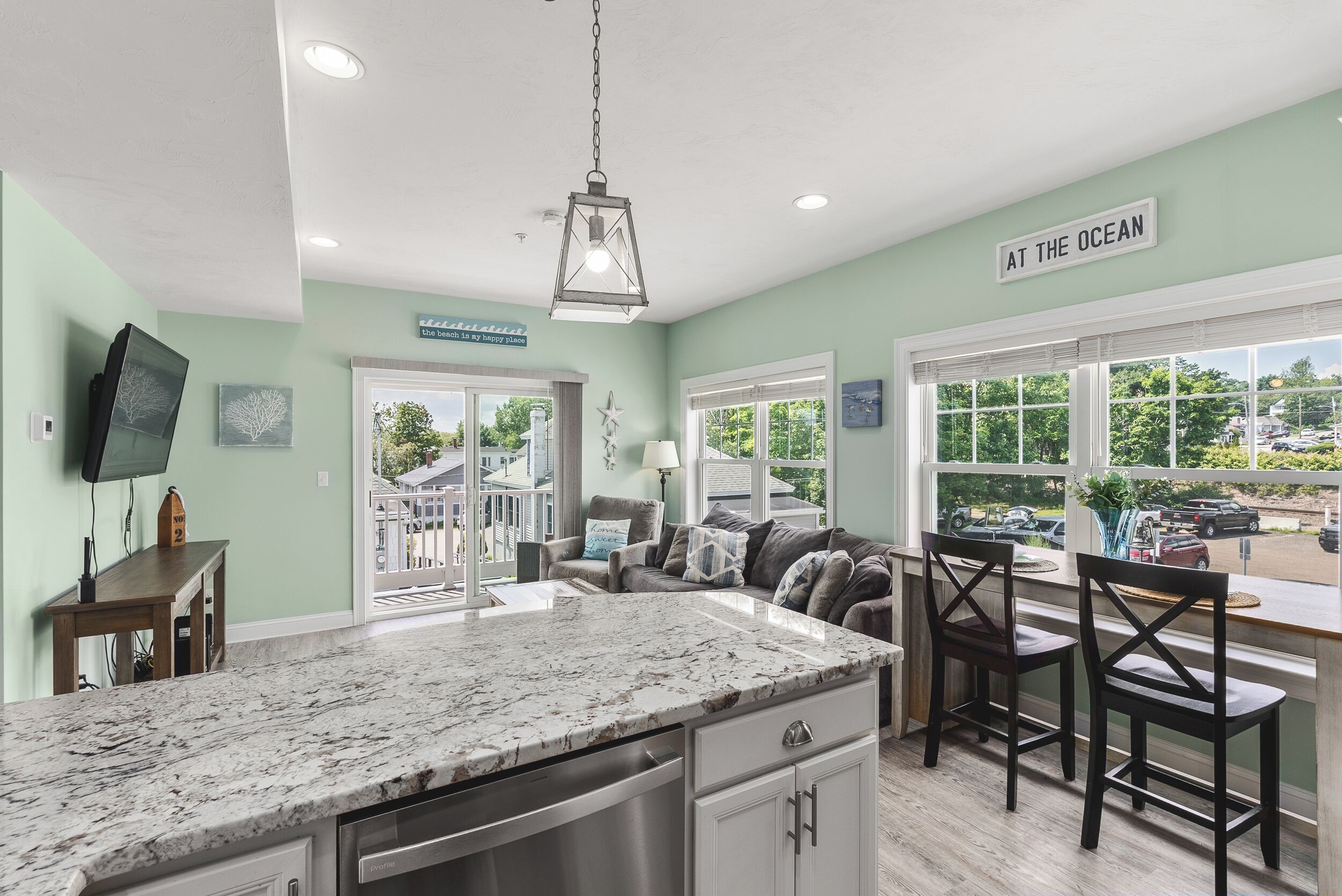 A kitchen with a large island, a dining area with chairs, a television mounted on the wall, and a sign that reads 'AT THE OCEAN'.