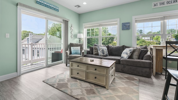 A living room with a sofa, a coffee table, and a chair. There are two windows with blinds, a ceiling fan, and a view of the outside.