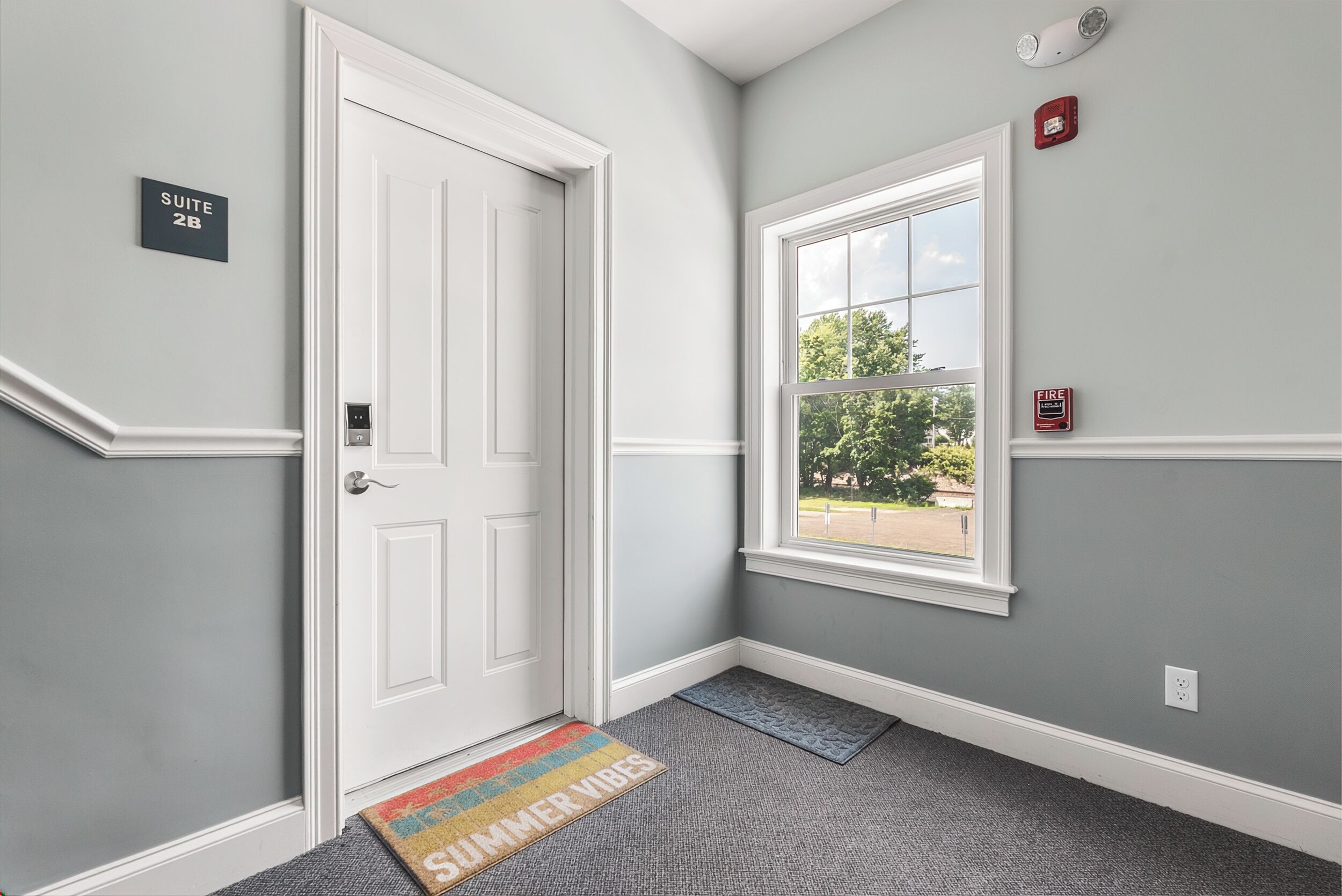 A hallway with a white door, a window with a view, and a fire alarm on the wall.
