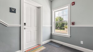 A hallway with a white door, a window with a view, and a fire alarm on the wall.