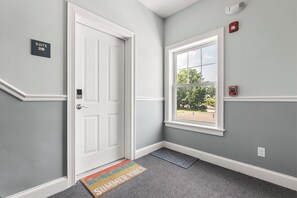 Un couloir avec une porte blanche, une fenêtre donnant sur un paysage agréable et un détecteur d’incendie au mur.