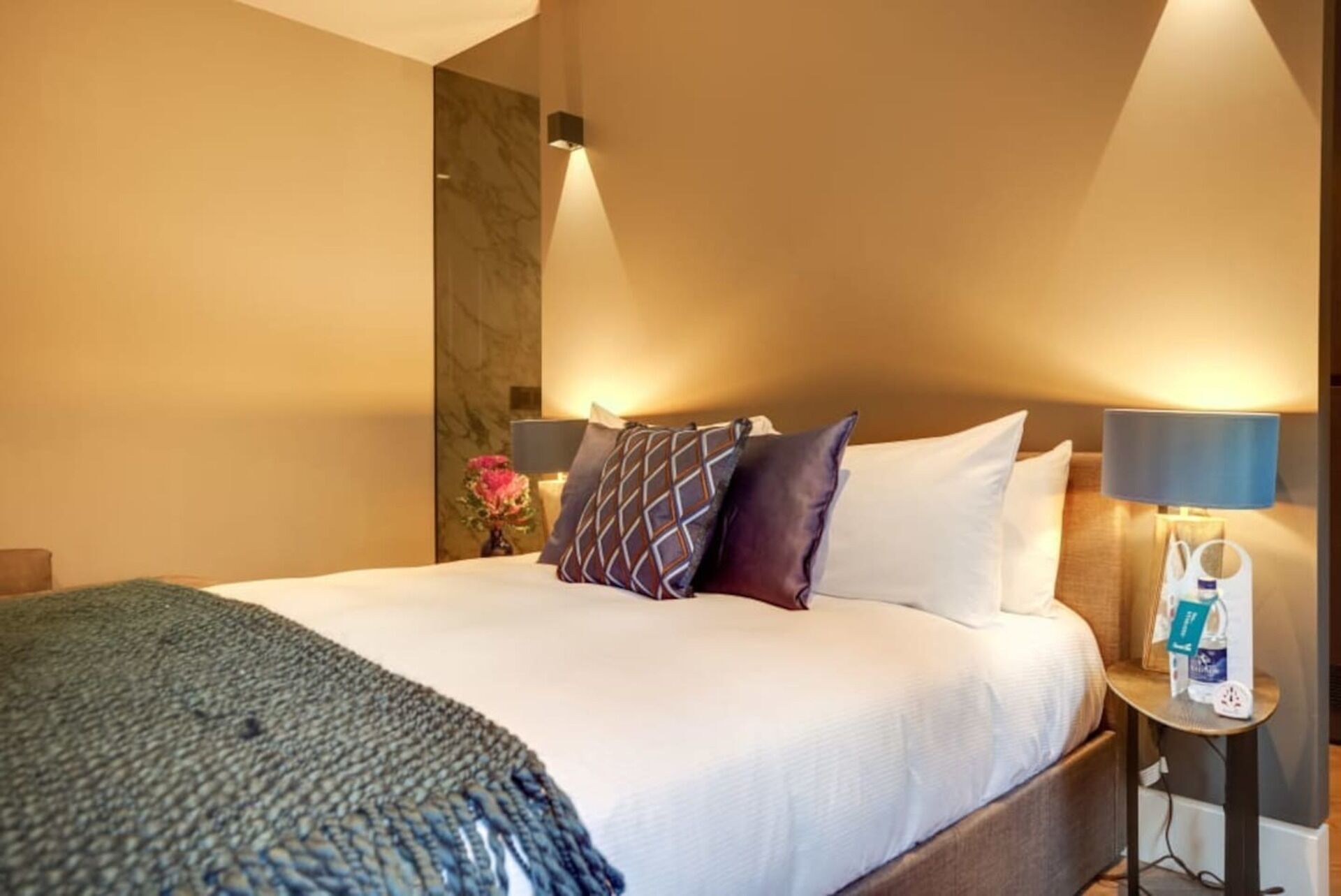 A hotel room with a large bed, a nightstand with a lamp, and a wall-mounted light fixture.