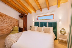 A bedroom with a large bed, wooden beams, a stone wall, a window, and a green chair.