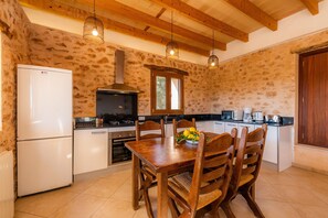 A kitchen with stone walls, wooden ceiling beams, a white refrigerator, a stove, and a dining table with chairs.