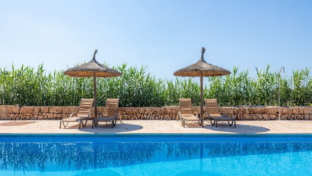 Piscina con sillas de descanso y sombrillas de paja, rodeada por un muro de piedra y vegetación exuberante.