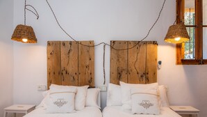 Un lit double avec une literie blanche et des coussins décoratifs, une tête de lit en bois, deux tables de chevet et des lampes suspendues en osier.