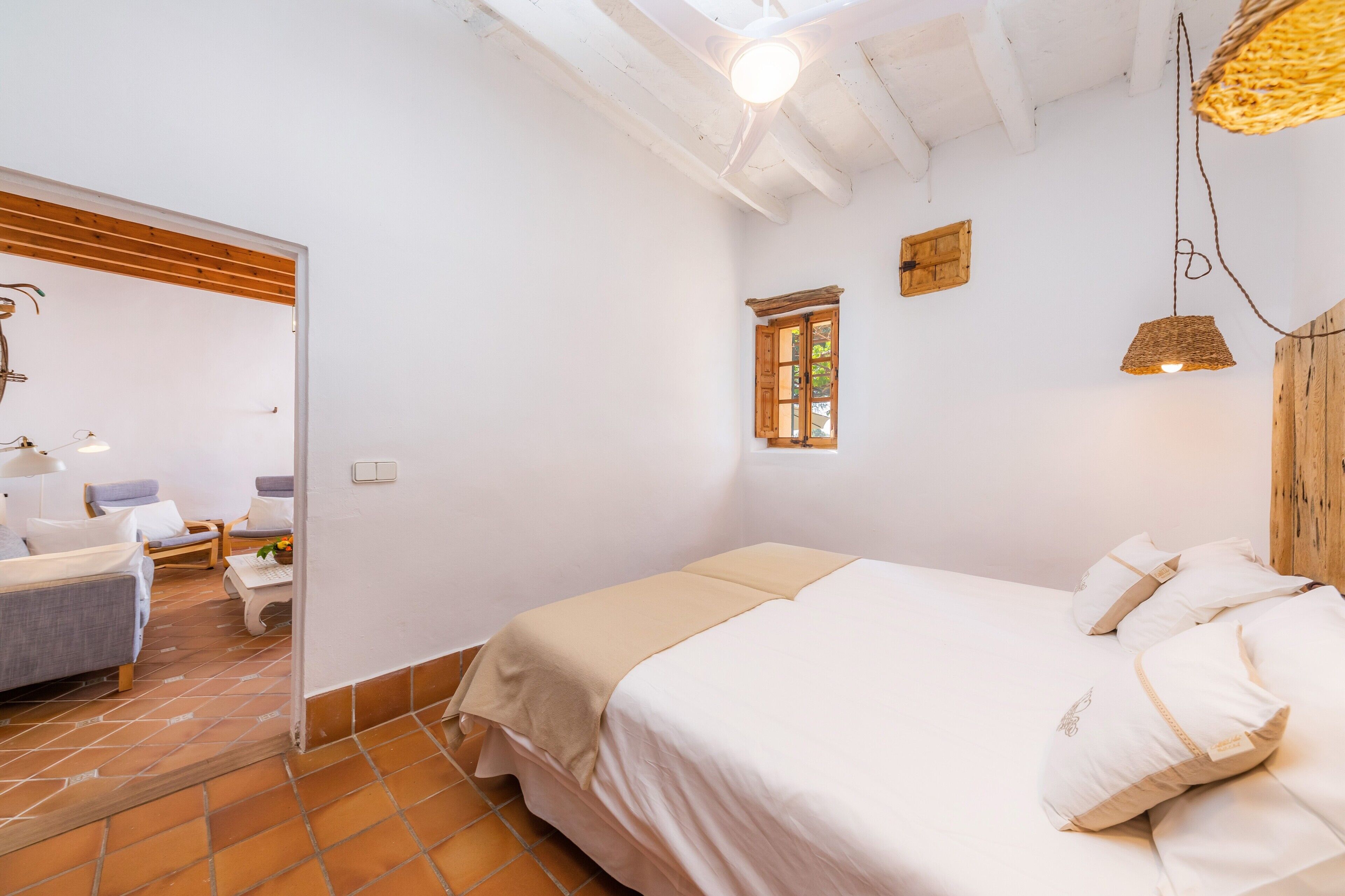 A bedroom with a bed, a small window, a hanging light fixture, and a wooden ceiling beam.