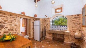 Une cuisine rustique aux murs de pierre, une table en bois agrémentée d’un centre de table composé de fruits, un réfrigérateur et une fenêtre donnant sur un paysage verdoyant.