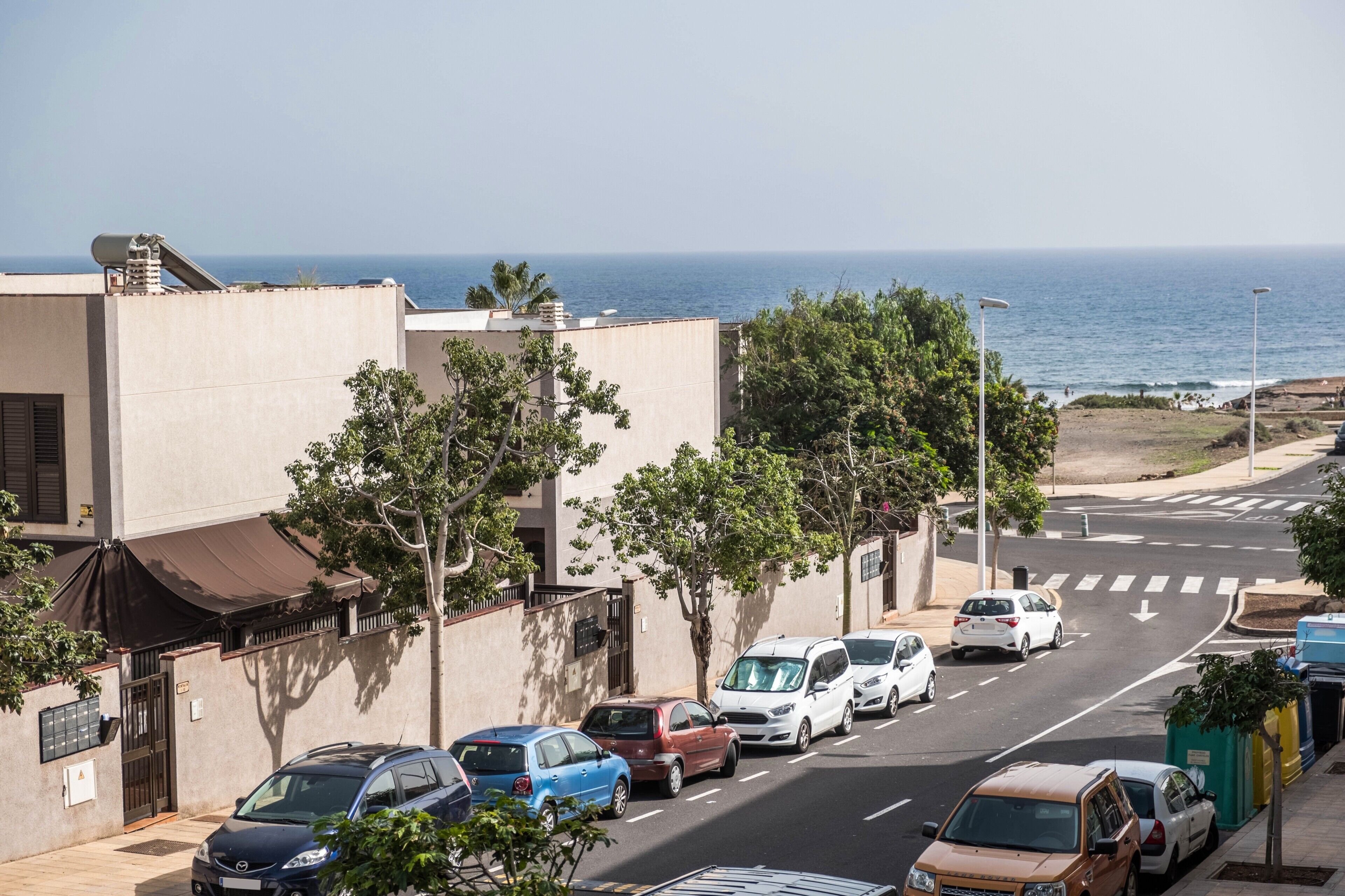 A coastal street with parked cars, a clear view of the ocean, and a building with a flat facade.