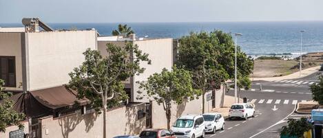 A coastal street with parked cars, a clear view of the ocean, and a building with a flat facade.