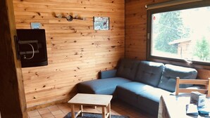 Een knus houten interieur met een grijze bank, een kleine houten tafel en een raam met uitzicht op bomen.