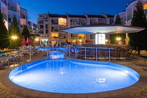 Una piscina con zona central hundida, rodeada de sillas de descanso y sombrillas, con una barra y edificios de varios pisos al fondo.