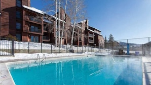Una piscina azul cristalina junto a un edificio con varios balcones y suelo cubierto de nieve.