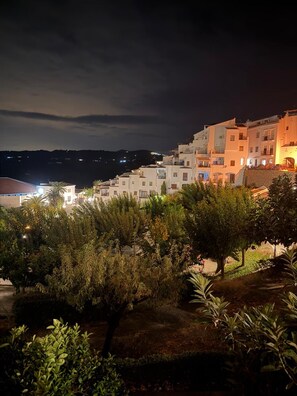 Vista nocturna de un pueblo costero con edificios blancos, una calle bien iluminada y abundante vegetación.
