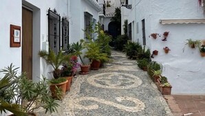 Une ruelle étroite pavée, bordée de bâtiments blancs dotés de balcons, de plantes en pot et surplombée d’un ciel dégagé.